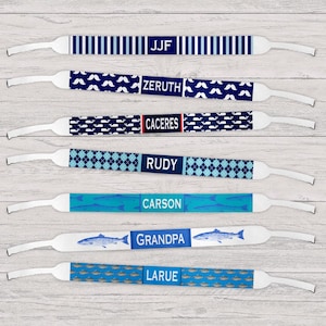 May include: A collection of personalized sunglass straps with various patterns and names. Designs include navy blue stripes, white mustaches, whales, argyle, fish, and small fish. Names include JJF, ZERUTH, CACERES, RUDY, CARSON, GRANDPA, and LARUE.
