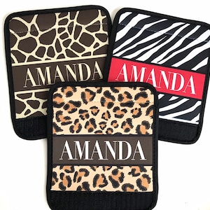 May include: Three personalized fabric wristlets with different animal print designs. The wristlets are black with a black strap and feature the name "Amanda" in white lettering. The designs include a brown and tan giraffe print, a black and white zebra print, and a brown and tan leopard print.