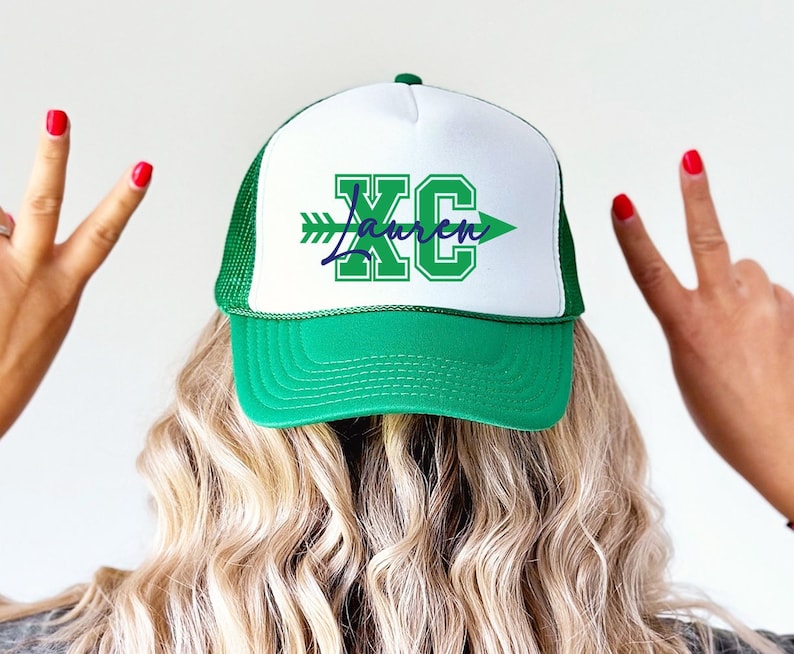 May include: A green and white trucker hat with the text "XC Lauren" in green and navy blue. The hat has a green brim and mesh back, with a white front panel. The text includes an arrow design.