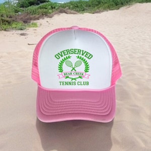 May include: Pink and white trucker hat with a green logo that says "Overserved Bear Creek Tennis Club" with two tennis rackets crossed in the middle.