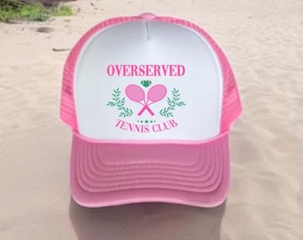 May include: A pink and white trucker hat with the text "OVERSERVED TENNIS CLUB" in pink. The hat features a pink brim and mesh back, with a white front panel displaying a tennis racket graphic.