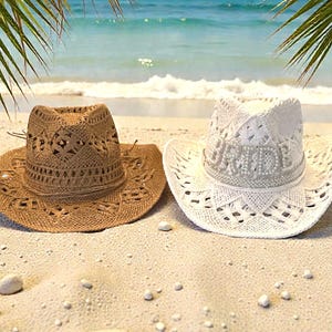 K&ouml;nnte beinhalten: Zwei Cowboyh&uuml;te an einem Sandstrand. Einer ist braun, der andere wei&szlig; mit dem Wort "BRIDE" in Perlen. Beide H&uuml;te haben eine breite Krempe und aufwendige Lochmuster, ideal als Sonnenschutz.