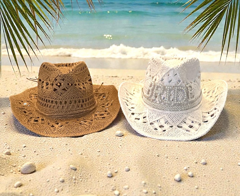 K&ouml;nnte beinhalten: Zwei Cowboyh&uuml;te an einem Sandstrand. Einer ist braun, der andere wei&szlig; mit dem Wort "BRIDE" in Perlen. Beide H&uuml;te haben eine breite Krempe und aufwendige Lochmuster, ideal als Sonnenschutz.