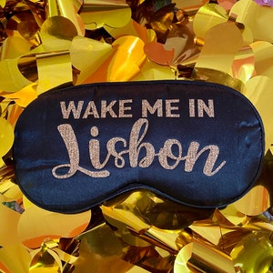 May include: Black sleep mask with gold glitter lettering that says "Wake Me In Lisbon". The mask is surrounded by gold foil confetti.
