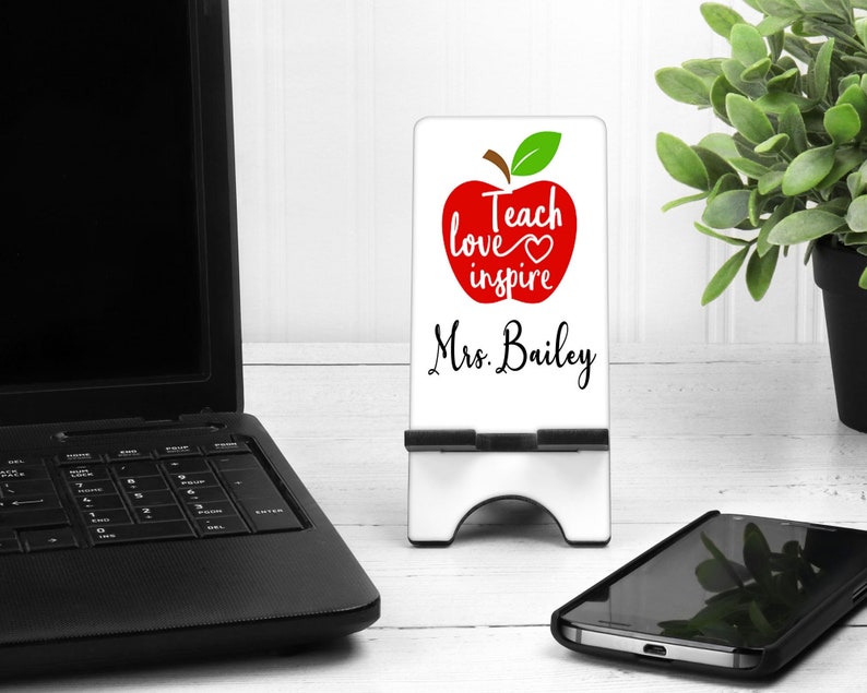 Puede incluir: Un soporte blanco para tel&eacute;fono con un dise&ntilde;o de manzana roja y las palabras "Teach love inspire" y "Mrs. Bailey" en escritura negra. El soporte est&aacute; sobre una superficie de madera blanca, con un port&aacute;til y un smartphone cerca.