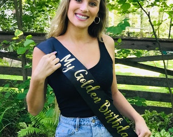 May include: A woman wearing a black tank top and blue jeans is holding a black sash with the gold text "My Golden Birthday" on it.