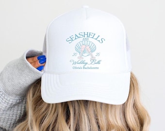 May include: White trucker hat with a blue and pink seashell and champagne glasses design. The text reads "Seashells and Wedding Bells Olivia's Bachelorette".