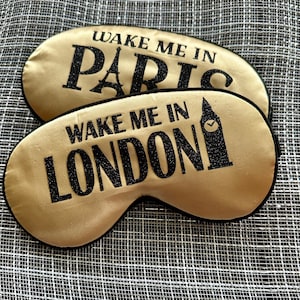 May include: Two gold satin sleep masks with black trim. The top mask says "Wake Me In Paris" and the bottom mask says "Wake Me In London".