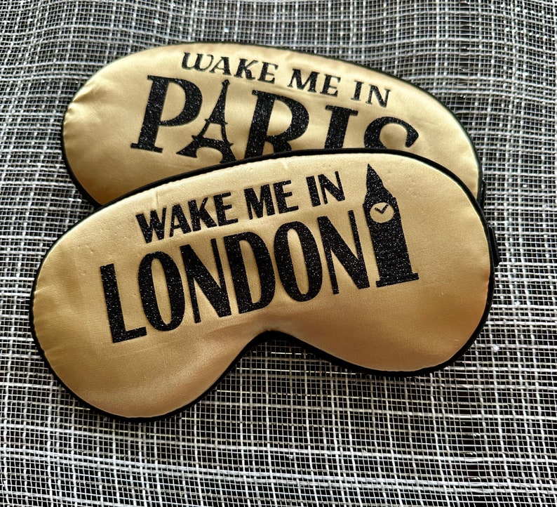 May include: Two gold satin sleep masks with black trim. The top mask says "Wake Me In Paris" and the bottom mask says "Wake Me In London".