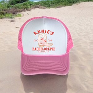May include: Pink trucker hat with a white front panel featuring the text "Annie's 20 24 Bachelorette Austin, TX" and a pair of pink cowboy boots with red accents.