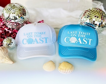 May include: Two trucker hats with a white seashell and the text "Last Toast on the Coast" in blue. One hat is white with blue text and the other is blue with white text.