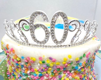 May include: A silver tiara with rhinestones spelling out the number "60" on top of a white cake with colorful sprinkles.