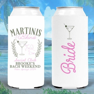 May include: Two white can coolers with a pink and gray design. One cooler says "Martini's & Bikinis" with a martini glass and a wreath. The other cooler says "Bride" in pink cursive.