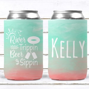 May include: Two teal and pink can coolers with white text. One cooler says "River Trippin Beer Sippin" with a sun, birds, a life preserver, and two beer cans. The other cooler says "Kelly".