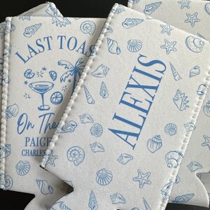 May include: White foam can coolers featuring a blue seashell and starfish pattern. One cooler displays the text LAST TOAST, a cocktail glass, a palm tree, and the names PAIGE and CHARLES. Another cooler features the name ALEXIS in blue text.