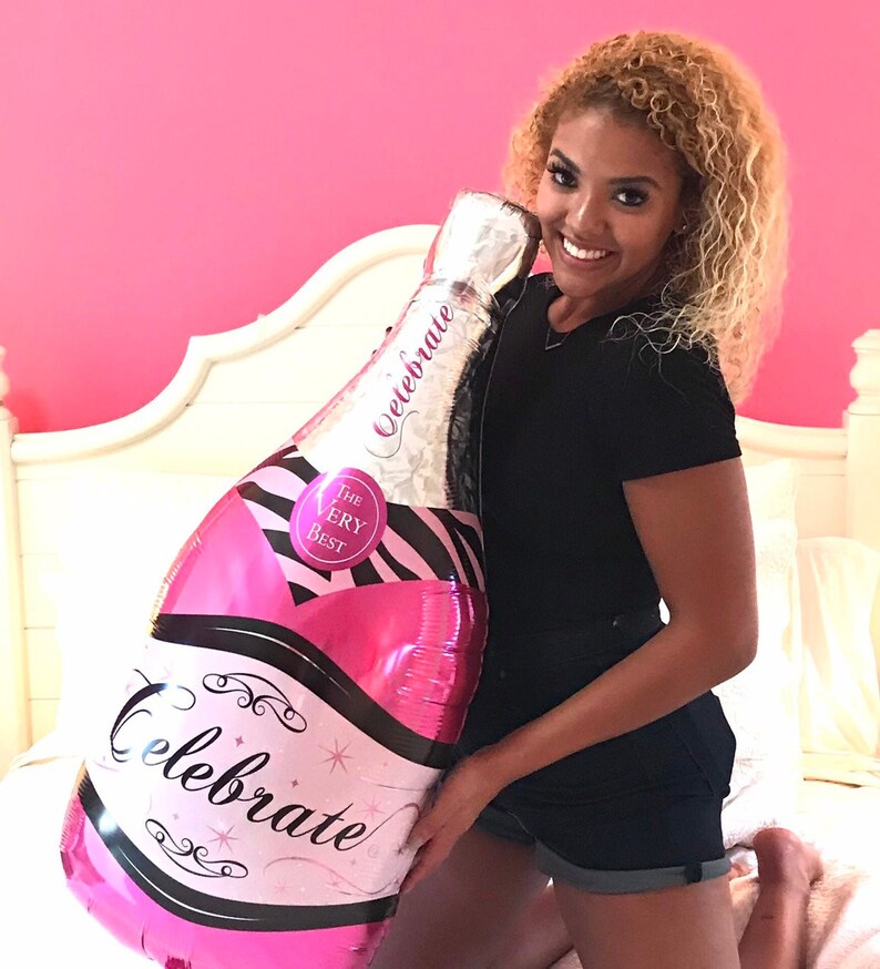 May include: A pink and black foil balloon shaped like a champagne bottle with the words "Celebrate" and "The Very Best" printed on it.