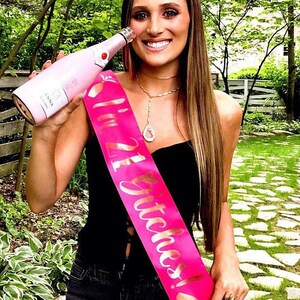 May include: A woman wearing a black strapless dress with a pink sash that says "21 Stitches!" She is holding a pink bottle of champagne.