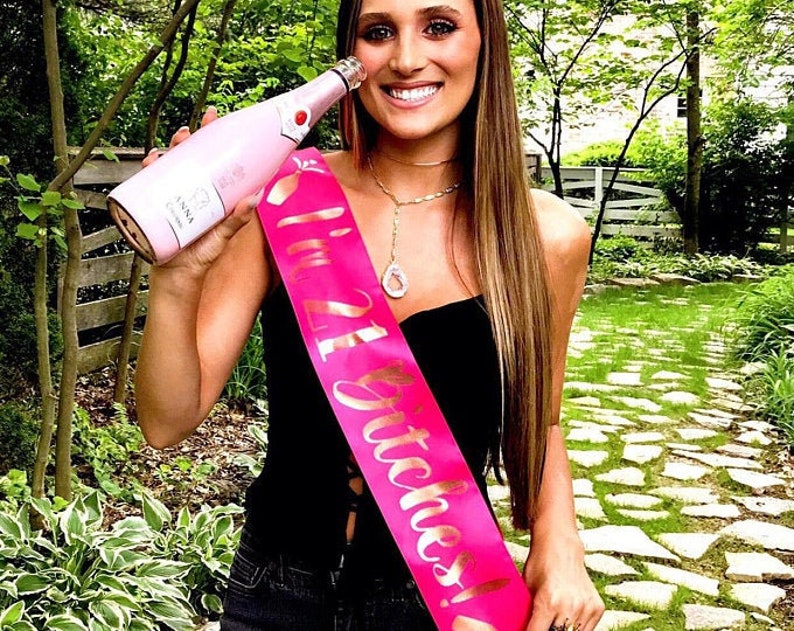 May include: A woman wearing a black strapless dress with a pink sash that says "21 Stitches!" She is holding a pink bottle of champagne.