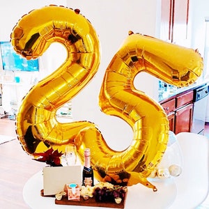 May include: Gold number balloons in the shape of 25, a bottle of champagne, a glass of champagne, grapes, and a cheese board on a white table.