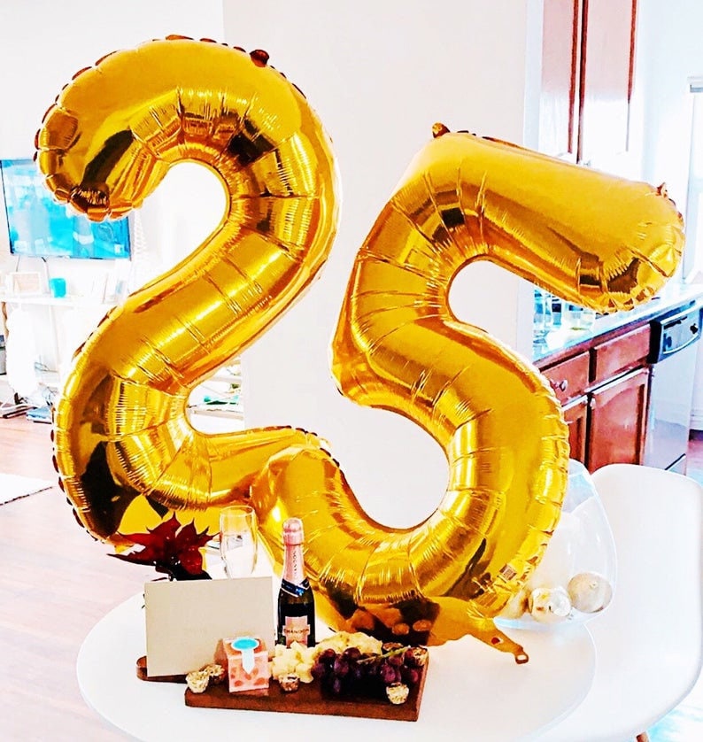 May include: Gold number balloons in the shape of 25, a bottle of champagne, a glass of champagne, grapes, and a cheese board on a white table.