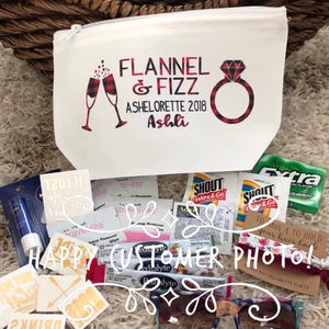 May include: A white cosmetic bag with a zipper, printed with "FLANNEL & FIZZ ASHELORETTE 2018 Ashli" in red and black. The bag is surrounded by small packets, sunglasses, and a hair tie, likely for a bachelorette party.