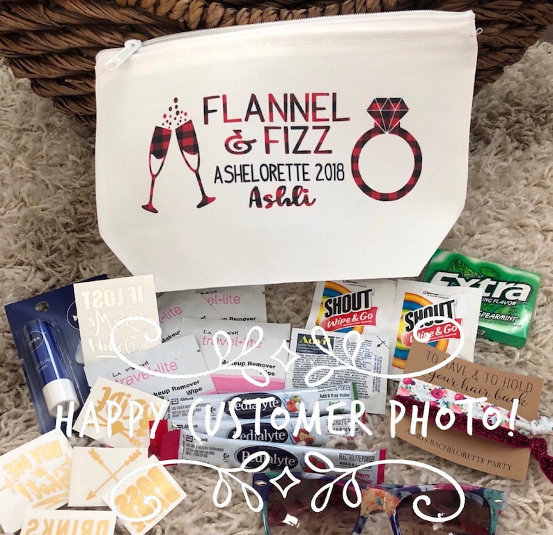 May include: A white cosmetic bag with a zipper, printed with "FLANNEL & FIZZ ASHELORETTE 2018 Ashli" in red and black. The bag is surrounded by small packets, sunglasses, and a hair tie, likely for a bachelorette party.