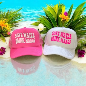 May include: Two trucker hats with the text "SAVE WATER DRINK MARGS". One hat is bright pink with white text, the other is white with pink text. Both hats have a mesh back and a curved brim. The hats are on a sandy surface with tropical plants in the background.