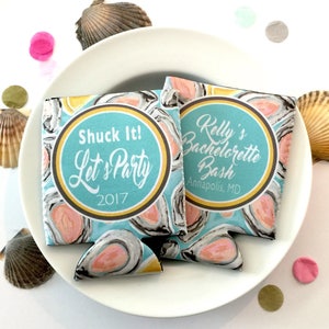 May include: Two blue and white can coolers with oyster shell designs and the text "Shuck It! Let's Party 2017" and "Kelly's Bachelorette Bash Annapolis, MD". The coolers are on a white plate with seashells and confetti.