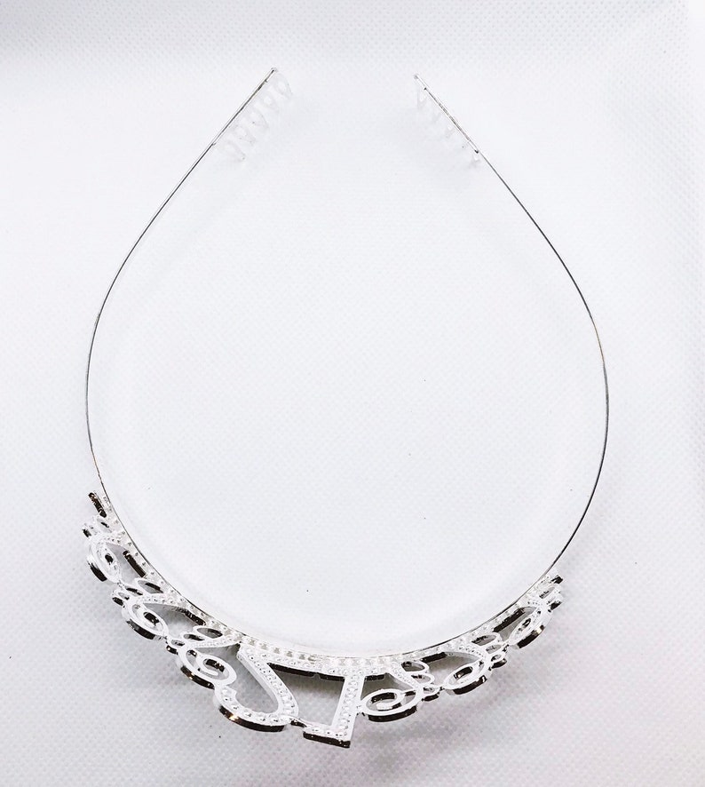 May include: A silver tiara with a decorative design at the base. The tiara has a clear plastic band for securing it to the head. The design includes the letters "J" and "S".