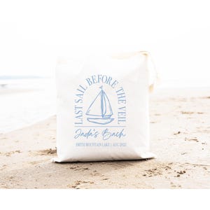 May include: A white canvas tote bag featuring a light blue graphic of a sailboat centred within an arch. The text reads LAST SAIL BEFORE THE VEIL, Jada's Bach, and SMITH MOUNTAIN LAKE | AUG 2025 in a mix of serif and script fonts.