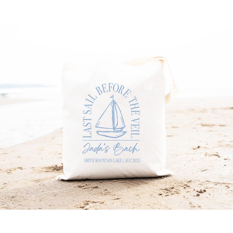 May include: A white canvas tote bag featuring a light blue graphic of a sailboat centred within an arch. The text reads LAST SAIL BEFORE THE VEIL, Jada's Bach, and SMITH MOUNTAIN LAKE | AUG 2025 in a mix of serif and script fonts.