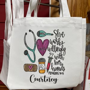 May include: White canvas tote bag featuring a pink heart, stethoscope, thermometer, syringe, bandage, and pill bottle. The text reads "She works willingly with her hands Proverbs 31:13" and "Courtney".