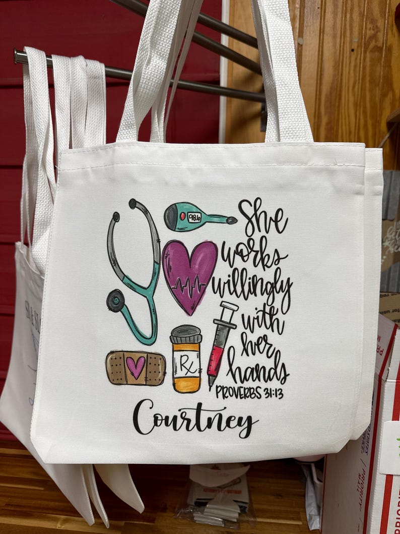 May include: White canvas tote bag featuring a pink heart, stethoscope, thermometer, syringe, bandage, and pill bottle. The text reads "She works willingly with her hands Proverbs 31:13" and "Courtney".