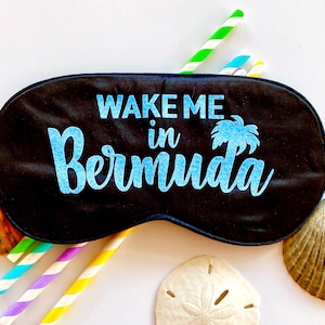 May include: Black sleep mask with the text "WAKE ME in Bermuda" in blue. The mask is surrounded by colorful straws, seashells, and a sand dollar on a white background. The mask has an elastic band.