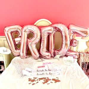 May include: Rose gold foil balloons spelling "BRIDE" are the focal point of this image, set against a pink backdrop. Additional decorations include a "bride to be" sash, cowboy hats, and confetti, creating a festive atmosphere.
