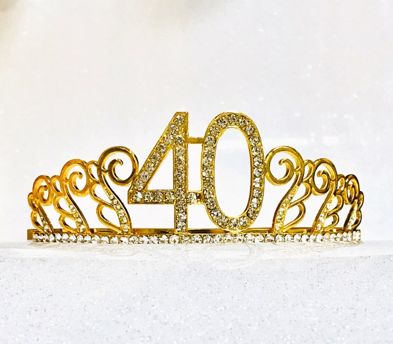 May include: A gold-colored tiara with the number 40 in the center, encrusted with rhinestones. The tiara has an ornate, filigree design with additional rhinestones along the base. Perfect for a 40th birthday celebration.