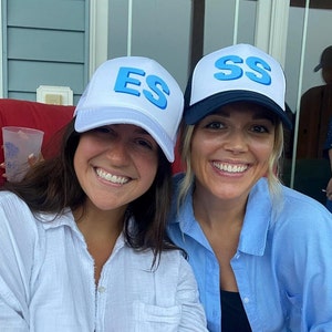 May include: Two women wearing white and black trucker hats with blue lettering. The hats say "ES" and "SS". The women are smiling and looking at the camera.