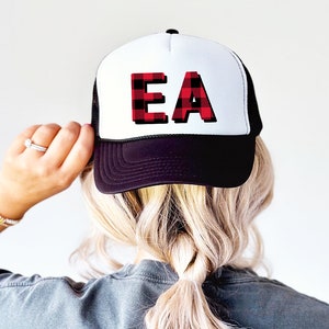May include: A black and white trucker hat with a red and black buffalo plaid "EA" design on the front.
