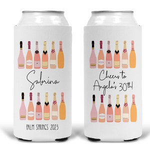 May include: Two white can coolers with pink champagne bottle designs. One cooler says "Sabrina, Palm Springs 2023" and the other says "Cheers to Angela's 30th!"