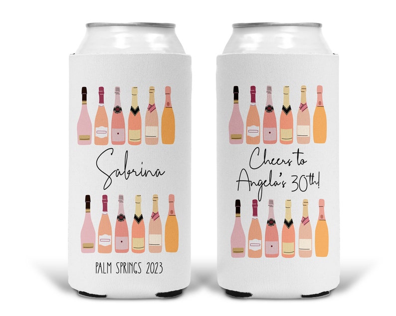 May include: Two white can coolers with pink champagne bottle designs. One cooler says "Sabrina, Palm Springs 2023" and the other says "Cheers to Angela's 30th!"