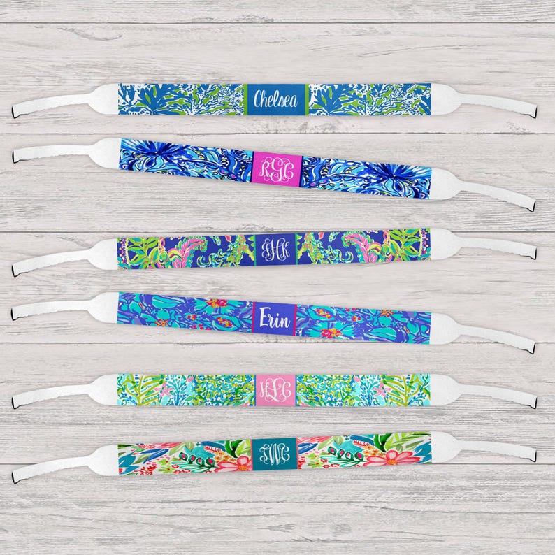 May include: Eyeglass straps with colorful floral patterns in blue, green, and pink. Each strap features a personalized name or initials on a rectangular label. The straps have white adjustable ends.