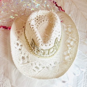 May include: White crochet cowboy hat with a beaded trim that says "Bride" in gold lettering.