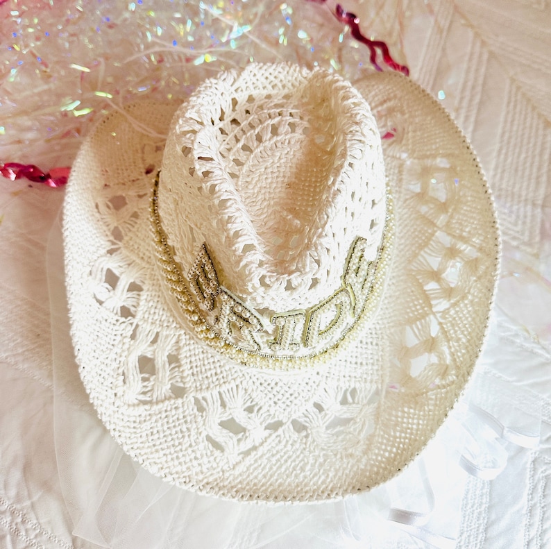 May include: White crochet cowboy hat with a beaded trim that says "Bride" in gold lettering.