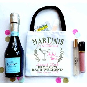 May include: White tote bag with a black handle and the text "Martinis & Bikinis Social Club Brooke's Bach Weekend Palm Springs 2024" printed on it. The bag is filled with a bottle of prosecco, a small bottle of lotion, and a small bottle of lip gloss.