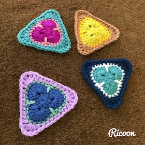 May include: Four crocheted triangular shapes in different colours and patterns. The shapes are made with a variety of coloured yarns, including pink, yellow, blue, and green. The shapes are arranged in a square pattern on a brown background.