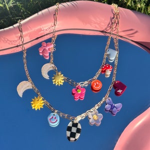 May include: Two gold chains with various charms, including a smiley face, a sun, a flower, a mushroom, a heart, a butterfly, a crescent moon, and a checkered oval. The charms are made of colorful plastic and are hanging from the chains.