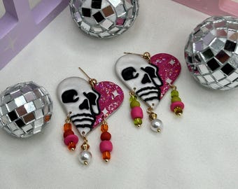 May include: Heart-shaped earrings with a skull design, featuring a white base and a pink glitter accent. The earrings have dangling beads in shades of pink, orange, green, and white, with gold-colored accents. Disco balls are in the background.