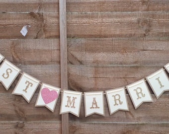May include: A banner that reads "JUST MARRIED" with individual flag-shaped panels. The flags are off-white with gold glitter lettering and a pink glitter heart. The banner is strung on twine and set against a wooden fence.