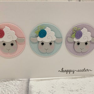 Op de afbeelding: Een witte paaskaart met drie schapenkoppen in pastelkleurige cirkels met bloemendetails. De schapen hebben witte wol en grijze gezichten. De woorden "happy easter" staan in zwarte letters onderaan de kaart.