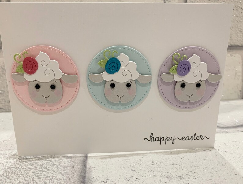 Op de afbeelding: Een witte paaskaart met drie schapenkoppen in pastelkleurige cirkels met bloemendetails. De schapen hebben witte wol en grijze gezichten. De woorden "happy easter" staan in zwarte letters onderaan de kaart.